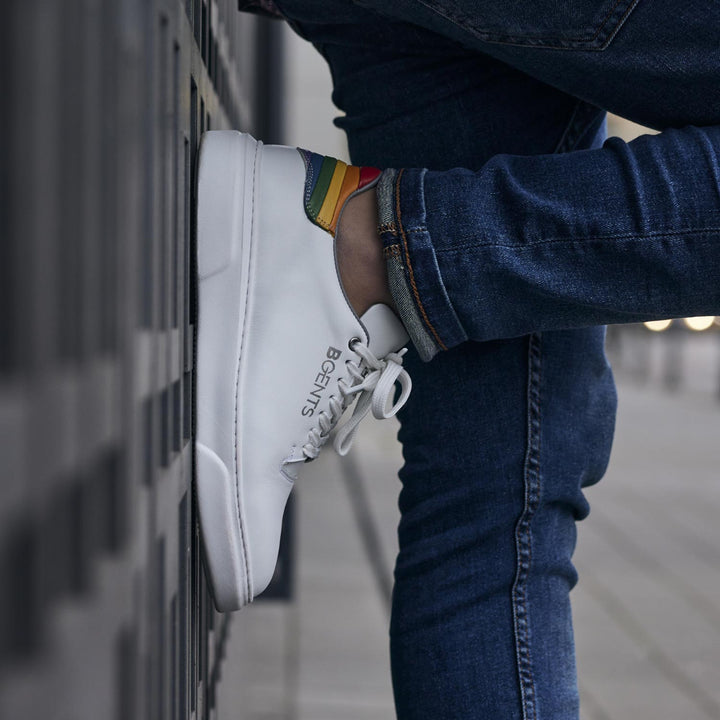  Weißer Pride Sneaker mit Regenbogen Farben an der Fersenkappe von BGENTS an der Wand gelehnt
